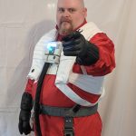 The writer dressed as a Resistance Pilot from Star Wars: The Last Jedi. Bald male, goatee, wearing a reddish jumpsuit, with a white life preserver-looking vest over it. He's pointing at the camera wearing black gloves.